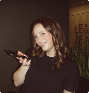 A young woman with wavy brown hair and a black shirt is smiling and holding a phone. She stands indoors against a dark background with green plants and part of a sign visible behind her.